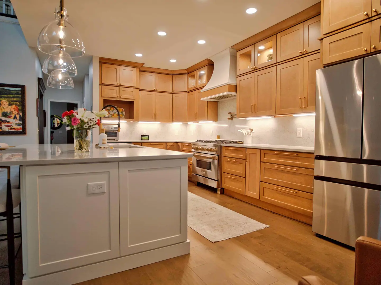 Open kitchen with a white island, maple cabinets with lit glass-front uppers and a stainless refrigerator