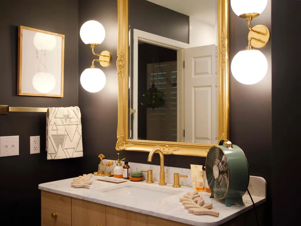 Black-walled bathroom with a gilded mirror, globe sconces, brass fixtures and a marble-topped oak vanity