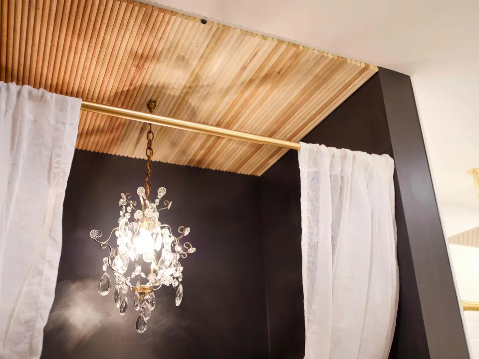 Bath alcove with a slatted wood ceiling, black walls, brass curtain rod and crystal chandelier