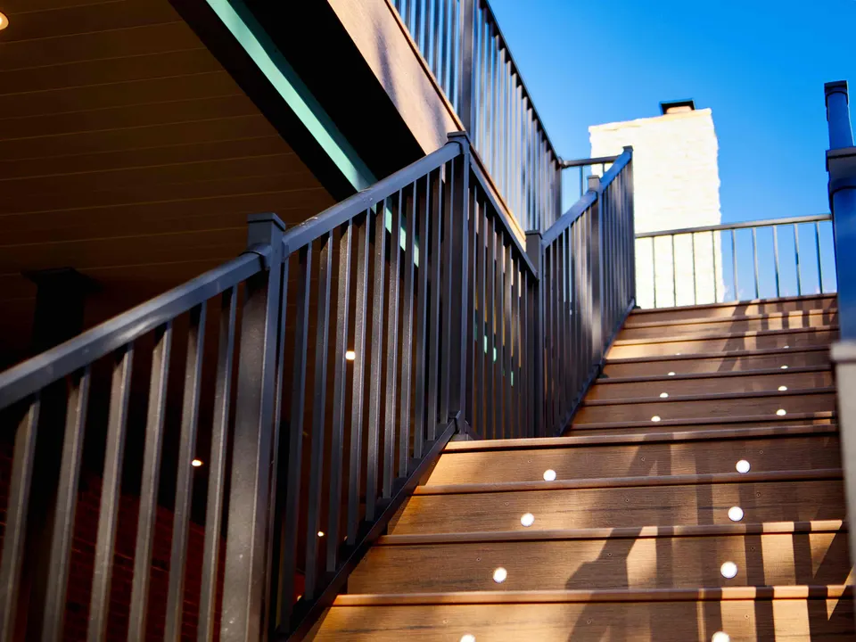 Composite deck stairs with built-in step lights and black railings beside a painted brick chimney