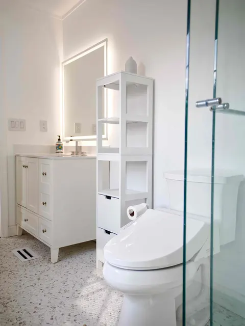 White bathroom with a shaker vanity, backlit mirror, open linen shelving and a smart toilet on a terrazzo-style floor