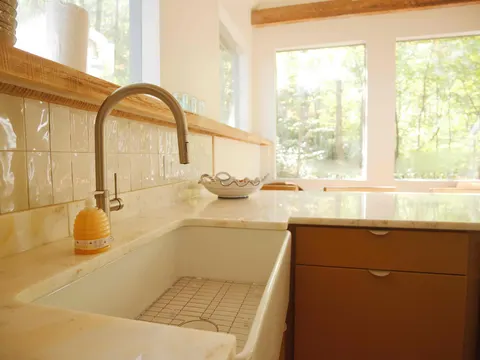 Fireclay farmhouse sink with a brushed gold faucet and glazed tile backsplash under a wall of windows